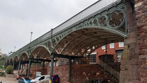 Iron Bridge in Exeter. It has large green-coloured iron girders and white iron underneath. A couple of brick walls are also underneath the bridge along with two cars and a white van which are parked up.