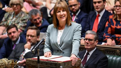 Rachel Reeves in a lightly coloured suit, pointing at her notes in a binder at the despatch box. Her party colleagues surround her, with Sir Keir Starmer sat closest to the right of the shot.
