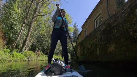 Sarah stands on her paddleboard, navigating her way down the river. The picture is taken from a miniature camera at the tip of her board, looking upwards at her.