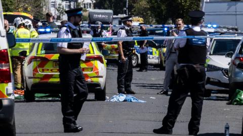 Police officers man a cordon as forensics staff in white suits and plain clothes detectives gather in Southport in the immediate aftermath of the attack on Monday, 29 July. A number of police vehicles including cars and vans can be seen inside the cordon.