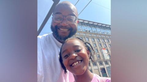 Anthony Ojo and his daughter. Anthony is bald and is wearing glasses and a white t-shirt. His daughter is wearing a pink t-shirt and has braids. Both are smiling to camera.