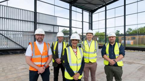 A group of four men and one woman stand in the construction area of a new building. A steel frame and roof can be seen in the background. One man is wearing an orange high-vis jacket, with the woman and three men wearing yellow high-vis jackets.