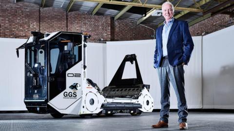 David is wearing a mid-blue blazer over a light blue shirt and light grey trousers. He is standing in a warehouse and in front of an autonomous baggage handling vehicle, made by his firm.