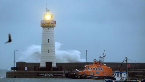 A wide shot of boats in front of a pier with a lighthouse on it. A wave can be seen striking the lighthouse from behind. The sky is grey. A bird can be seen.
