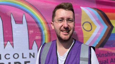 Arran Hart, wearing purple high vis and smiling in front of colourful Lincoln Pride banners