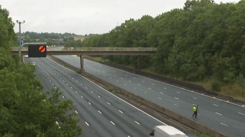 The northbound and southbound carriageway of the M1 motorway with no traffic. A man is stood on one side of the road wearing a fluorescent yellow jacket.