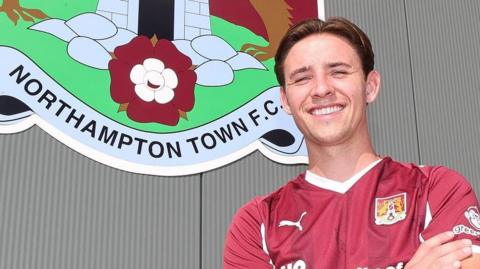 Terry Taylor standing in front of the Northampton Town club crest at Sixfields