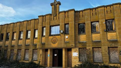 The image shows a two-storey building constructed from yellow stone, with rows of windows lining the façade. A stone cross sits at the top of the building, set against a bright blue sky. Many of the ground-floor windows are boarded up, and security cameras and warning signs are in place to deter trespassers.