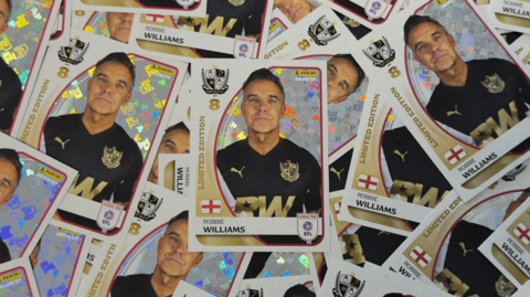 A scattered arrangement of Panini football stickers, all of which feature Robbie Williams, a man wearing a black football top with brand logos and the letters RW in gold type on the front.
