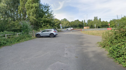 A rural car park with two cars parked a little distance away from each other. Trees and foliage appear in both the left foreground and the background. A red parking gate is also seen in the background with a number of traffic signs.