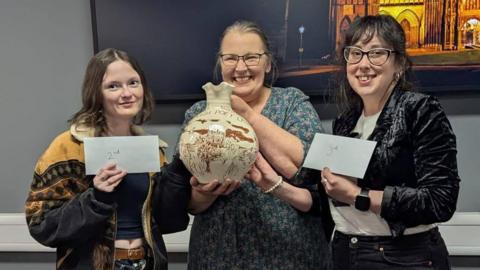 Three finalists, with Hilary Steele in the middle, smile and hold their prizes. Hilary is holding a jug. The second and third-placed poets, both women, are holding envelopes.