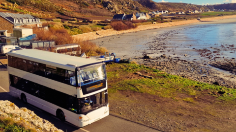 A white double-decker bus drives along a narrow coastal road beside a rocky, partly wet beach. The shoreline curves gently with shallow waves, while a row of small houses and cottages sits at the base of low, rugged hills covered in brown and green vegetation. The sky is partly cloudy with warm sunlight casting soft shadows across the landscape.