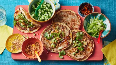 A red chopping board sits on a blue table cover. On top sits tortillas with a smashed layer of minced pork and bowls filled with garnishes. There is chopped cucumber, spring onions,  crispy onions, sesame seeds and a chilli-infused oil.
