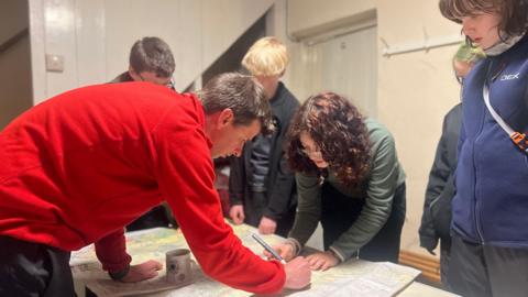 A team leader helping students draw a route on a large map on a table. There is a girl leaning over the table with a pen as two people beside her watch. 