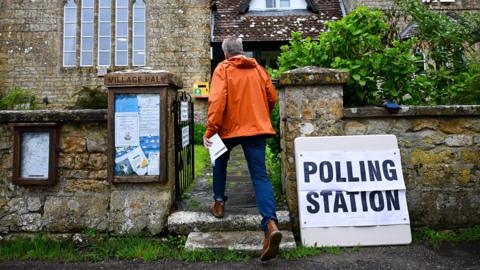 A man walking into a polling station in a rural church . He is seen from behind walking through a gate and wearing an orange coat 