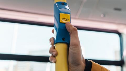 A generic shot inside a bus, showing a close up of a man's thumb that is about to press a "stop" button on a pole.