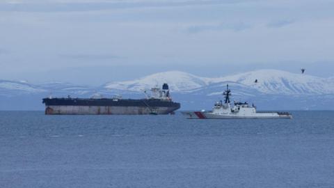 The tanker is a large ship with rusty marks on its hull. The upper part of its hull is painted blue and it has a white bridge and a single blue funnel. In the foreground is a white US Coast Guard vessel. There are snow-covered hills in the background.