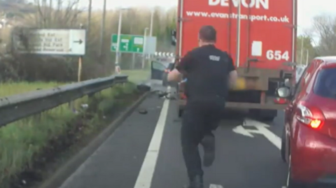 A police officer runs on the road towards a red HGV, and in front of it a black car. The door of the car can be seen open, with a sign in front of it warning of an approaching roundabout.