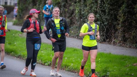 Mark Davies in the centre of the picture wearing running gear, flanked by two support workers, also wearing brightly coloured running kit.  All three are running along a street as part of the Conwy half marathon.