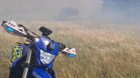 A blue, yellow and white motorbike parked up in an dry straw field with smoke in the background.
