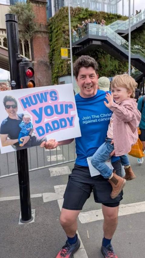 A man in athletic clothes holds a small child while posing on a busy street during the Great Manchester Run, surrounded by crowds and barriers. The man carries a sign reading “HOW’S YOUR DADDY” with a photo, with a brick bridge, greenery-covered wall, and a staircase with onlookers in the background.