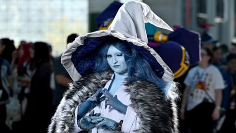 A fan of Elden Ring at last year's Comnic Con in New York dressed as Elden Ring's Ranni the Witch, in a grey gown, huge silver hat, fur cape and blue skin make-up, with an extra set of arms and hands and long, blue hair