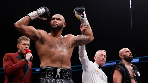 Frazer Clarke in the ring with his hands aloft after winning a fight. A ring announcer in a red jacket is on the left with a microphone, while a referee holds Clarke's left arm up. His opponent Bogdan Dinu looks dejected on the right