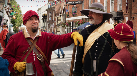A group of people dressed in 17th-Century attire gather on a cobbled street outside the George Hotel, taking part in a historical re-enactment. They are wearing wide-brimmed hats, doublets, and gloves, with one individual holding a metal tankard. Bunting and hanging flower baskets add to the festive atmosphere in this picturesque market town setting.