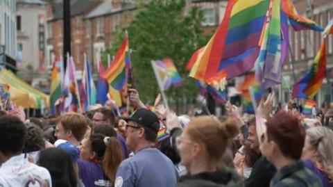 A crowd of people holding rainbow flags in a city street, standing in profile.