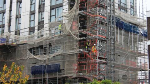 Builders work on a construction site in london. Three men can be seen climbing scaffolding while wearing fluorescent jackets and hard hats.