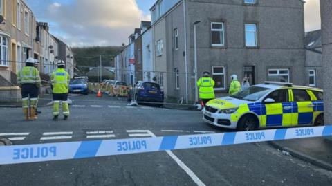 Officers in high-vis jackets stand on a street, which is partly fenced off. A police car and police tape are in the foreground.
