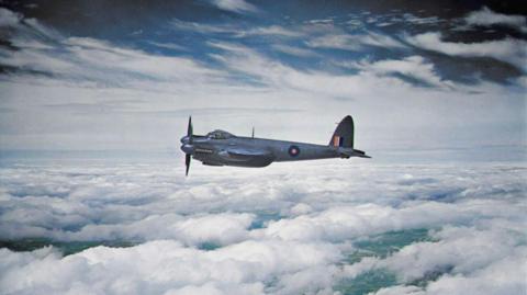 A colour picture taken of the RAF Mosquito in mid-flight from another aircraft in 1943. The plane is flying above the clouds and is a blue-grey colour with two propellers. 