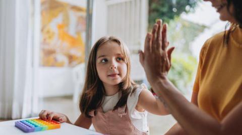 A mother and daughter play with a brightly coloured pop it fidgit toy at a table. The pair are sharing a high five, and the girl, who has shoulder-length brown hair, is wearing pink dungarees and white T-shirt. The mother is wearing a mustard top.