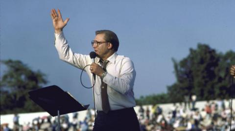 Jimmy Swaggart holds a microphone and gestures with his hand as he preaches to a large crowd outside during the 1980s.