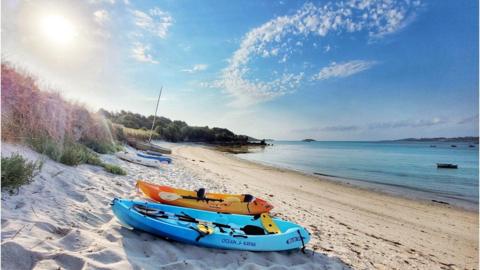 A blue and a yellow boat sit on white sand with blue see to the right and a wisp of white cloud in a blue sky