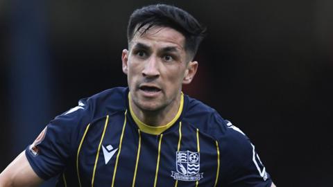 A photo of footballer Noor Husin playing for Southend United Football Club. Husin is looking to his right while running on the pitch with an open mouth, wearing a dark blue shirt with yellow strips and neck trim and the Southend badge and kit logo in white either side of his chest