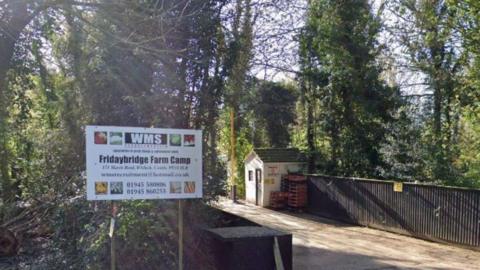 A view of the front entrance of a farm camp for agricultural workers. There is a sign with the name of the company - WMS - and the site name - Fridaybridge Farm Camp. There is a small road going across what looks like a bridge into the camp and a small wooden shed with pallets stacked against one side