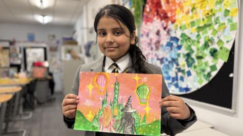 Maidah, who has black hair in a ponytail and wears her grey school uniform jacket and tie, holds up her winning artwork in an art classroom. It shows the backs of Elphaba and Glinda holding hands as they look on to the Emerald City, which has green towers and structures and hills sloping down on both sides. Two hot air balloons - one pink and the other green - glide above through a pinky-orange sunset.