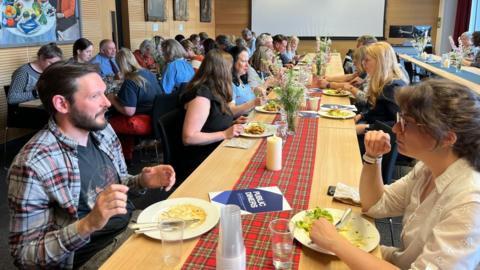 A group of people sitting at a long dinner table. The guests are sitting across from each other in a well lit room. The table features a red tartan centrepiece with false candles and flowers on it.