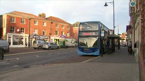 A bus is stood at a bus stop on the right next to a pavement where people are standing. On the left there are cars and vans parked on a small car park in front of shops.