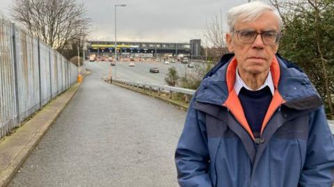 John McGoldrick is standing by a slip road to the Kingsway Tunnel in Wallasey. He is looking at the camera and is wearing a blue and orange raincoat with a navy blue sweater and blue and white striped shirt. He has grey hair and is wearing glasses. A number of cars can be seen queuing to go through the tunnel. 