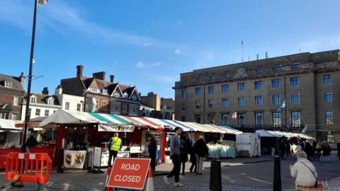 The Market Square on a sunny day with people visiting the outdoor stalls. They have different coloured tarp roofs and are in the shadow or the large Guildhall building. It is light bricked and towers over the square.