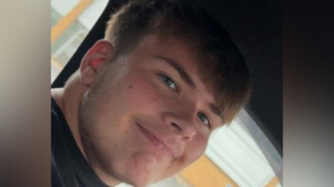 Jamie Nolan, a teenage boy, is sitting in the passenger seat of a car smiling at the camera. He has short brown hair, blue eyes and is wearing a black t-shirt. 