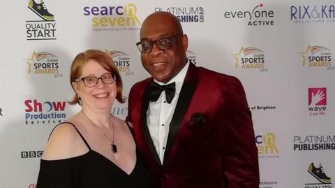 Eric and Mandi Douglin pose before a sponsor board at an awards event. Both are wearing formal attire.