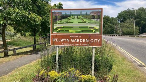 A sign featuring an illustration of a formal garden in a brown frame with white lettering saying "Welcome to Welwyn Garden City". There is a road running past the sign. There are shrubs around the bottom of the sign and trees on either side of the picture.