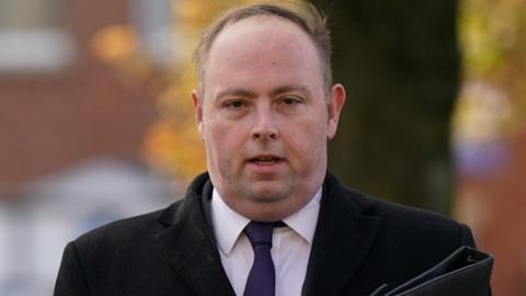 A man with short brown hair wearing black coat and tie