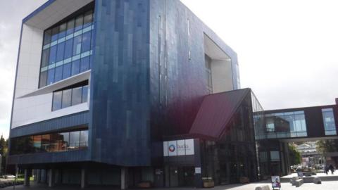 Buckinghamshire New University campus in High Wycombe. Part of the building is curved with blue panels and houses the university's library. There is a walkway connecting campus buildings and a white sign which reads BUCKS NEW UNIVERSITY.