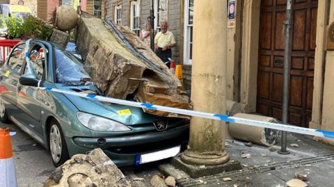 Car with smashed stones on top outside the pub. A large piece of masonry is resting on the bonnet and front window of the car