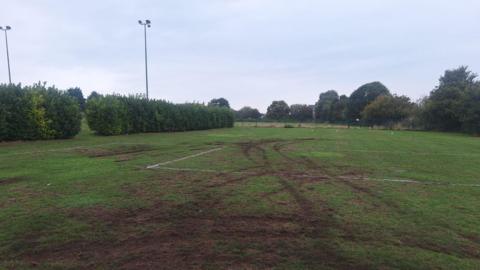 A sports field with areas of damage. Across the grass, there are deep tire tracks and several bare patches where the grass has been torn up, exposing the soil underneath. On the left side, tall hedges run along the edge of the field, and in the distance, trees form a natural border. 