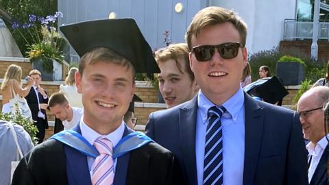Jack and Ben O'Sullivan pictured at Jack's graduation. Jack is 23 and Ben is 27. They are white men with short, mousey brown hair. Jack is dressed in black graduation robes and a mortarboard, which he wears over a blue suit with pale blue shirt and pink tie. Ben also wears a blue suit, pale blue shirt and navy blue tie, with dark framed sunglasses. They're pictured outside on a sunny day around other graduates in the background.